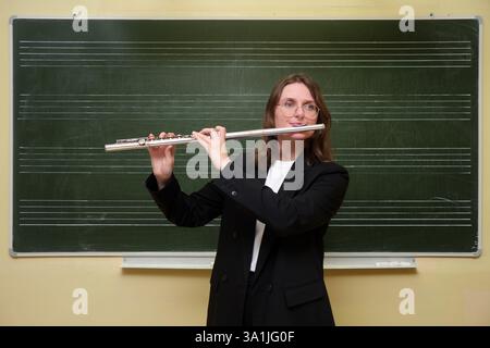 The smiling female teacher, a skilled flutist, stands by the blackboard in her classroom, happily playing a classic melody on her flute while her elem Stock Photo