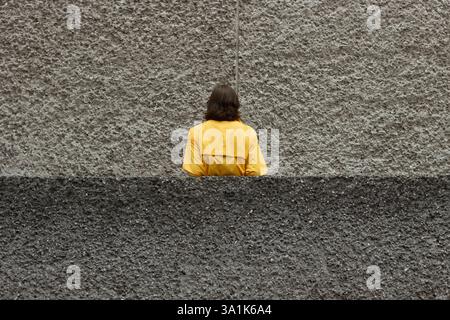 Woman in a bright yellow jacket stands with her back to the camera, leaning against a concrete balcony facade. The gritty, textured wall enhances the Stock Photo
