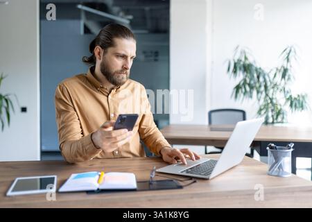 Bearded man works at a laptop while also using a mobile phone at his desk in a brightly lit modern office, looking focused. Stock Photo