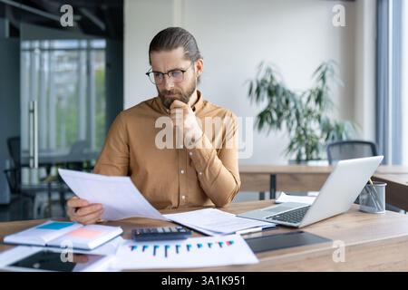 Bearded businessman in glasses examines documents at his desk with a laptop, calculator, and charts, thoughtfully analyzing information. Stock Photo