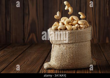 Cashew nuts are falling into burlap sack on rustic wooden table Stock Photo