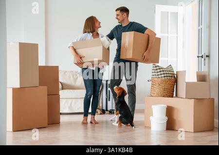 Walking, holding boxes and smiling. Young couple with dog are moving to new home. Stock Photo