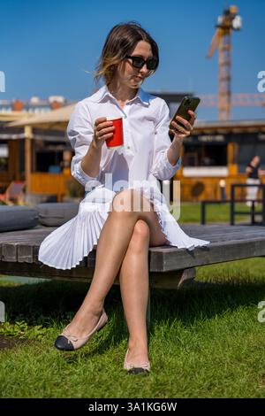 Woman in white dress wearing sunglasses and ballet flats, looking at smartphone while holding cup Stock Photo
