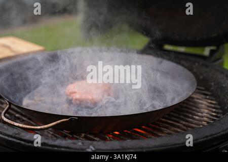 A large round wood-burning grill is roasting beef patty for burger. Fresh meat cutlet in a frying pan grill. Barbecue festival in the city park. Stock Photo