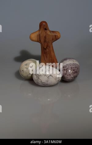 Three stone eggs in different colours, an olive wood cross standing above them & a plain white background. Symbolic of the Christian Easter festival. Stock Photo