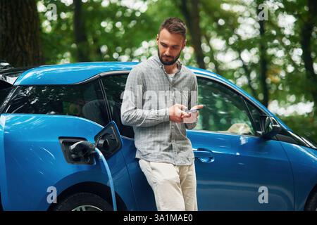 Wireless device, using smartphone. Handsome man is standing outdoors near electric automobile and waiting for charging. Stock Photo