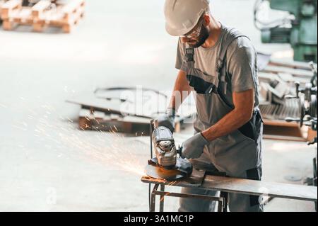 Angle grinder in hands. Young factory worker in grey uniform. Stock Photo