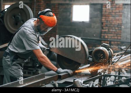 Side view, cutting the metal. Young factory worker in grey uniform. Stock Photo