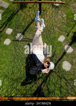 The image portrays a person relaxing on a net hammock outdoors. Stock Photo