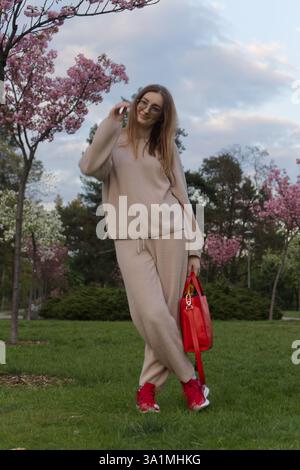 Girl stands in a park during early evening, dressed in a beige tracksuit and holding a red handbag. Stock Photo