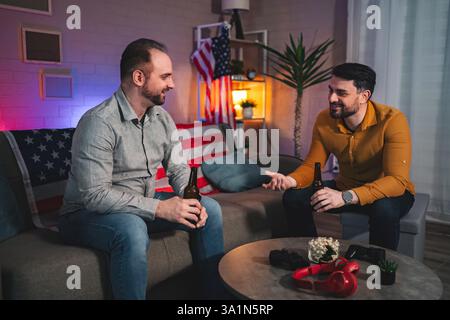 Two friends share stories and laughter over drinks in a cozy living room filled with warm ambiance and American decor Stock Photo