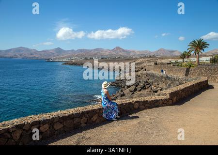 Woman holidaymaker sitting on the wall of the coastal path between Puerto del Carmen and Puerto Calero on the Spanish Canary island of Lanzarote Spain Stock Photo