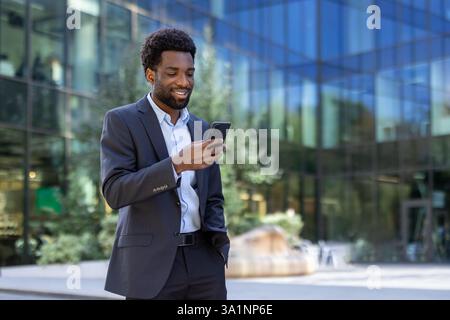 Successful businessman walking through the city with phone in hand. Joyful African American man in business suit using smartphone app, browsing social media, and typing text message. Stock Photo