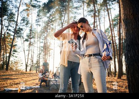 Two women are standing with smartphone. Group of friends are together in the forest. Stock Photo