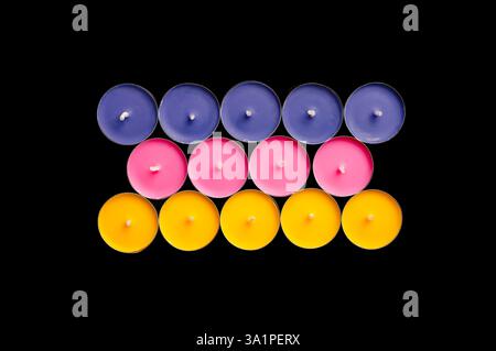 Tea lights in pink, yellow, and blue, arranged in a playful composition against a stark black backdrop. Stock Photo
