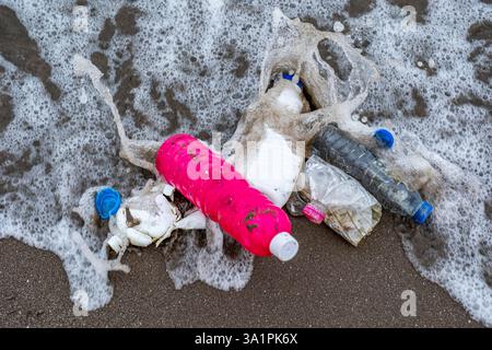 A bottle of pink shampoo is on the beach next to a pile of trash. The bottle is partially submerged in the water, and the trash is scattered around it Stock Photo