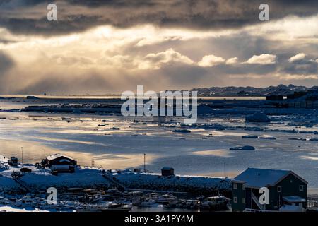 A serene winter landscape featuring a harbor filled with icebergs and boats. The sky is dramatic with dark clouds and rays of sunlight breaking throug Stock Photo
