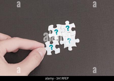 A hand holding a white puzzle piece with blue question mark, about to connect with three others. The puzzle rests on a dark textured surface Stock Photo