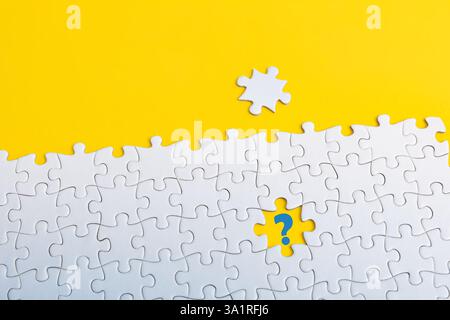 A white jigsaw puzzle on yellow background. A piece is missing in the center, with a blue question mark, a puzzle piece placed above Stock Photo
