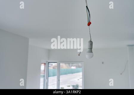 Single light bulb hanging from exposed electrical wires in unfinished interior with white walls and concrete flooring. Lighting in building under reno Stock Photo