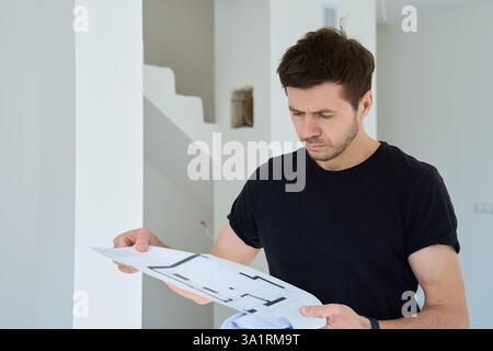 Man in black shirt examines architectural floor plan inside in unfinished apartment. Concept of real estate investment and renovation planning Stock Photo