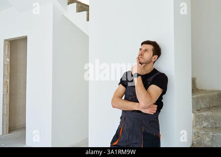 Thoughtful professional construction worker in black overalls standing in unfinished apartment. Man thinking about renovation plans in newly purchased Stock Photo