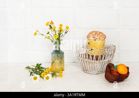Easter cake and colored Easter eggs in the wicker baskets with yellow Ranunculus asiaticus in the glass vase. Stock Photo