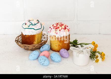 Sweet Easter cakes, colorful Easter eggs, a gift box and flowers with a white wall on the background. Stock Photo