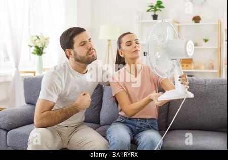 Unhappy young couple badly suffer with heat, using fan air for cooling or ventilation  Stock Photo