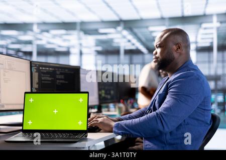 Server room programmer using green screen laptop to find firewall misconfigurations. African american man checking systems bottlenecks leading to sluggish data transfer rates using mockup notebook Stock Photo