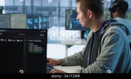 IT technician looking at PC screen, working in network security data center, ensuring optimal performance. Man supervising server hub, monitoring energy consumption across rigs, camera A Stock Photo