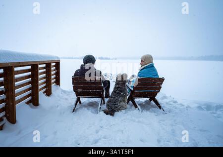 Couple with dog sitting on snowy lakeside enjoying winter scenery. Stock Photo