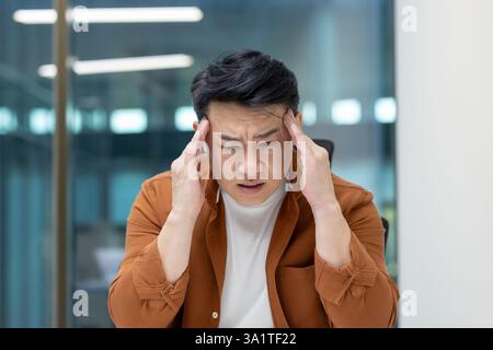Headache in office worker at workplace. Man suffering from migraine while working inside office. Stock Photo