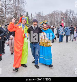 Yekaterinburg, Russia - February 26, 2025: Festive farewell to winter in city park. Cheerful man poses with jester and woman in folk attire with large Stock Photo