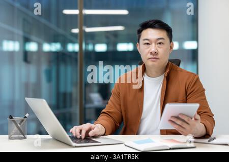 An Asian businessman is multitasking, working on his laptop while simultaneously using a tablet for a presentation in the office. Stock Photo