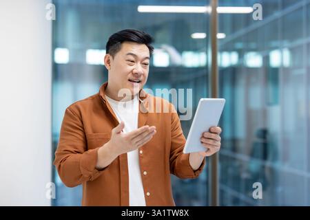 Successful Asian businessman talking to colleagues and partners using video call and tablet computer. Man standing near window inside company office. Stock Photo