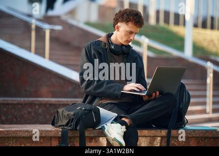 Guy is sitting and using laptop. Young student is outside the university, in black clothes. Stock Photo