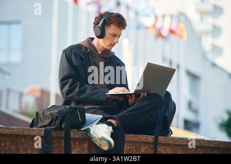 In headphones, using laptop. Young student is outside the university, in black clothes. Stock Photo