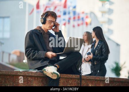 In headphones, using laptop. Young student is outside the university, in black clothes. Stock Photo