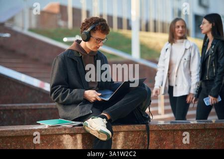 In headphones, using laptop. Young student is outside the university, in black clothes. Stock Photo