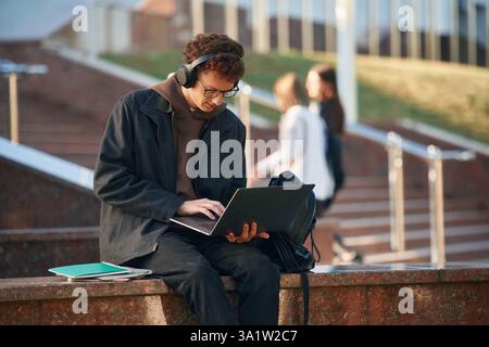 Guy is sitting and using laptop. Young student is outside the university, in black clothes. Stock Photo