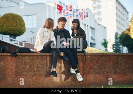 Sitting and using laptop. Three young students are outside the university outdoors. Stock Photo