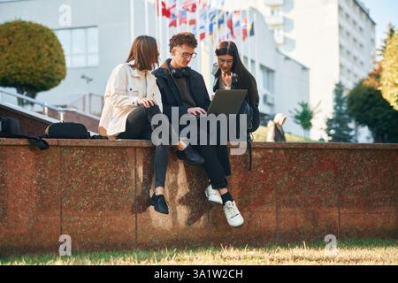 Sitting and using laptop. Three young students are outside the university outdoors. Stock Photo