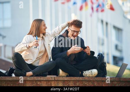 Smiling, having fun. Two young students are sitting outdoors against university. Stock Photo