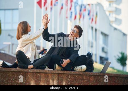 Giving high five, success. Two young students are sitting outdoors against university. Stock Photo