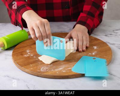 Child's hands using blue scraper to cut dough into pieces on wooden cutting board surrounded by rolling pin and other scrapers Stock Photo