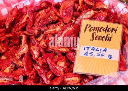 Close-up of sun-dried tomatoes with deep red tones and rough uneven surfaces piled in a plastic-lined bin with a label indicating origin and price at Stock Photo