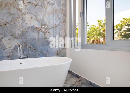 Bright and elegant modern bathroom interior featuring a sleek white freestanding bathtub positioned against a glossy marble wall and near large window Stock Photo