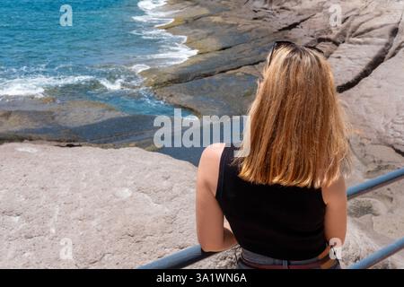 Rear view of a woman with shoulder-length blonde hair standing towards the ocean, young female leaning against a metal railing while gazing out Stock Photo