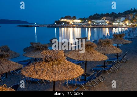 Umbrella lined white sand beach with fishing village behind at night, Finikounda, Messenia, Peloponnese, Greece, Europe Stock Photo